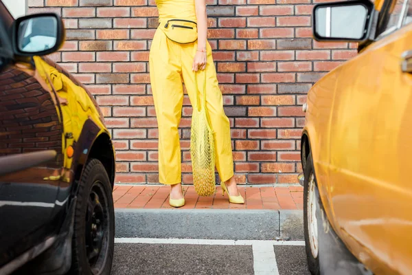 cropped shot of woman in yellow clothes with pineapple in string bag standing at parking in front of brick wall