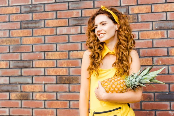 smiling young woman in yellow clothes holding pineapple in front of brick wall
