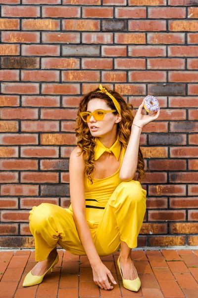 beautiful young woman in yellow clothes sitting in front of brick wall with donut in hand