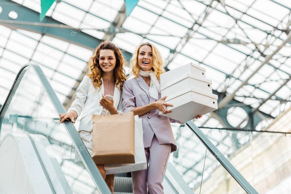 fashionable young women with shopping bags and boxes on escalator at mall
