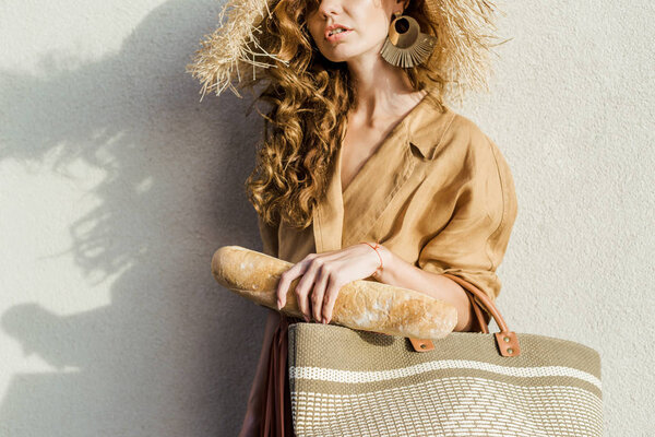 cropped shot of stylish woman in straw hat with baguette standing in front of white wall