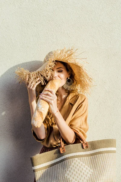 食べるバゲットの白い壁の前に立っている麦わら帽子の美しい若い女性 — ストック写真