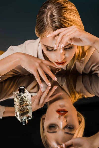 stylish blond woman at mirror surface with perfume on dark background