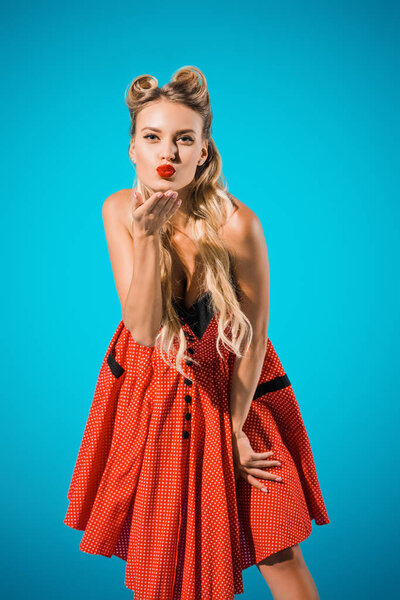 portrait of attractive pin up woman in retro style clothing blowing kiss on blue backdrop
