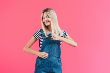 Genel olarak başparmak gösterilmesini ve pink izole kamera bakarak denim mutlu güzel kız