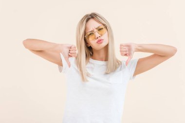 unsatisfied young woman in sunglasses showing thumbs down and looking at camera isolated on beige clipart