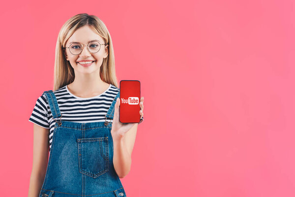 portrait of smiling woman in eyeglasses showing smartphone with youtube sign isolated on pink