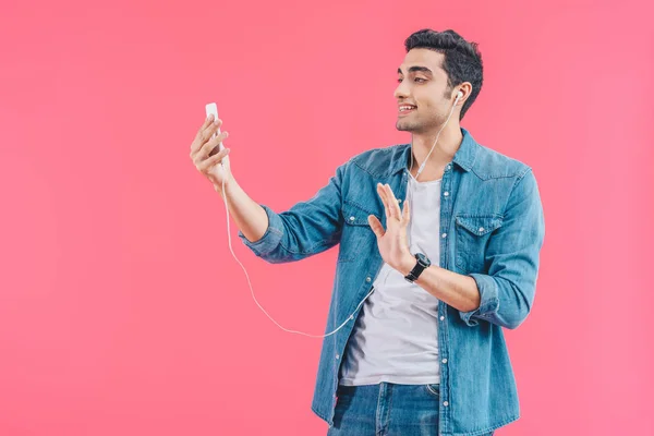 portrait of young man in earphones waving to smartphone isolated on pink