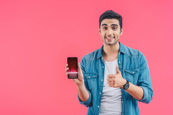 smiling young man doing thumb up gesture and showing smartphone with trading courses isolated on pink