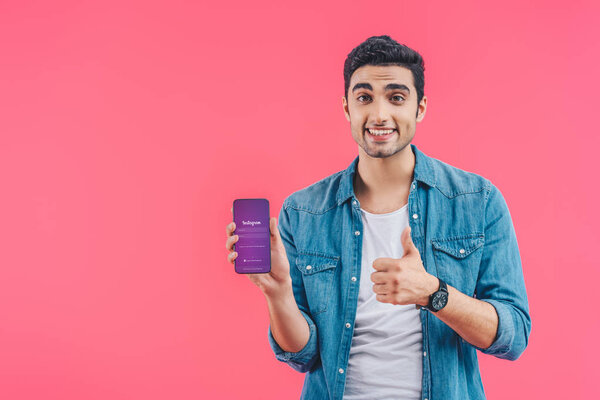 smiling young man doing thumb up gesture and showing smartphone with instagram website isolated on pink