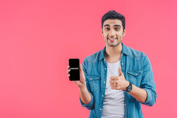 cheerful young man doing thumb up gesture and showing samsung smartphone isolated on pink