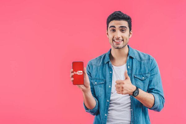 happy young man doing thumb up gesture and showing smartphone with youtube website isolated on pink