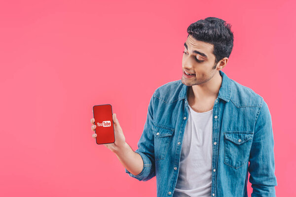 smiling young man showing smartphone with youtube website isolated on pink