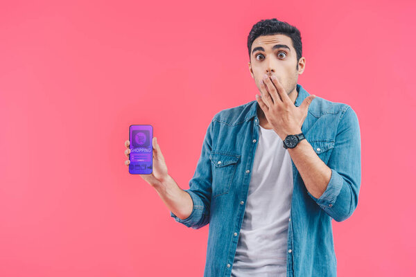 shocked young man covering mouth by hand and showing smartphone with shopping isolated on pink