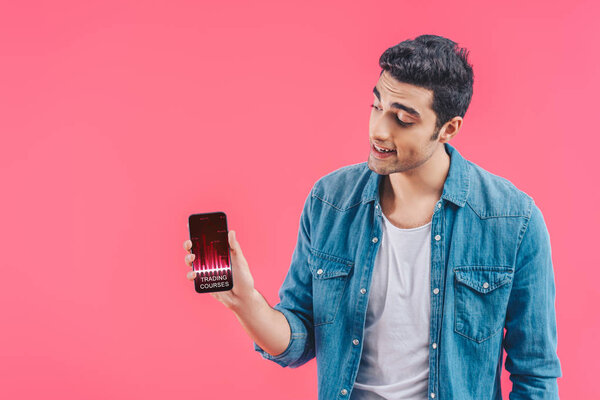 smiling young man showing smartphone with trading courses isolated on pink