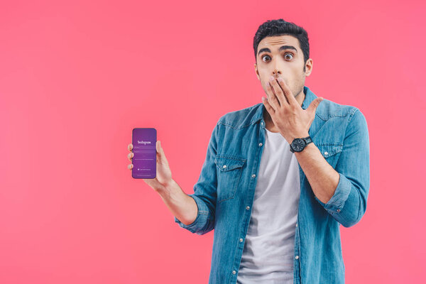 shocked young man covering mouth by hand and showing smartphone with instagram website isolated on pink