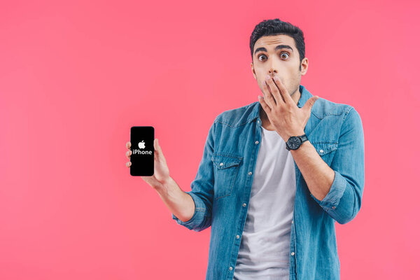 shocked young man covering mouth by hand and showing iphone smartphone isolated on pink