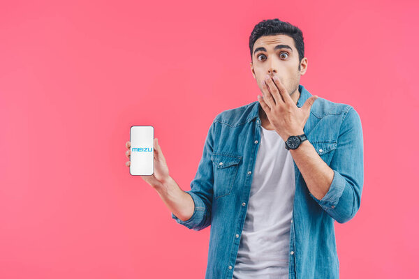 shocked young man covering mouth by hand and meizu smartphone isolated on pink