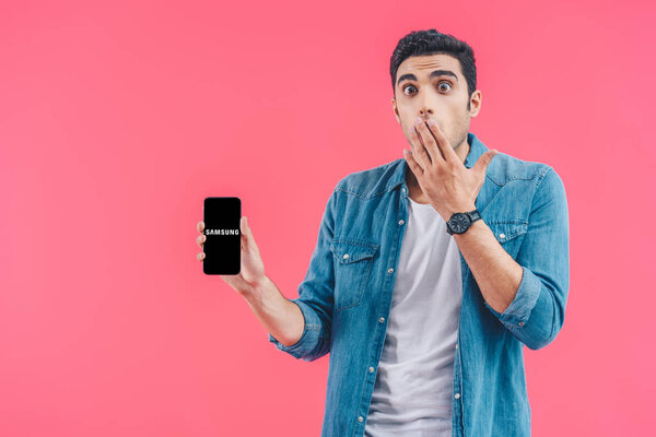 shocked young man covering mouth by hand and showing samsung smartphoneisolated on pink