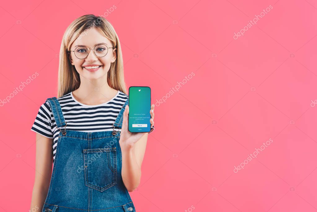 Portrait of smiling woman in eyeglasses showing smartphone with twitter logo isolated on pink