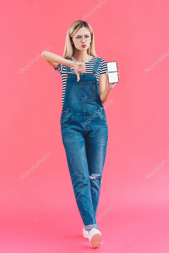 Young woman with smartphone with samsung logo showing thumb down on pink backdrop
