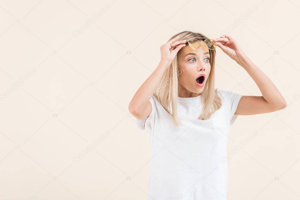 shocked blonde girl adjusting sunglasses and looking away isolated on beige