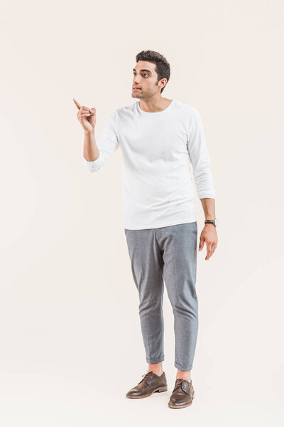 handsome young man looking away and pointing with finger isolated on beige  