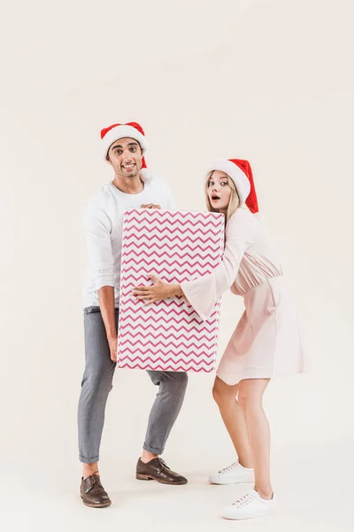 Young Couple Santa Hats Holding Big Christmas Present Looking Camera — Free Stock Photo