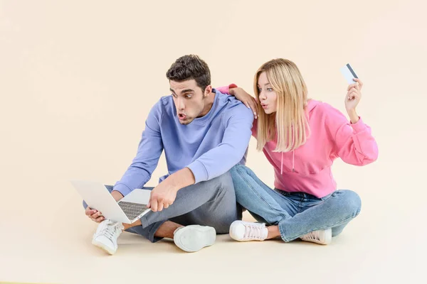 shocked young couple with credit card using laptop isolated on beige