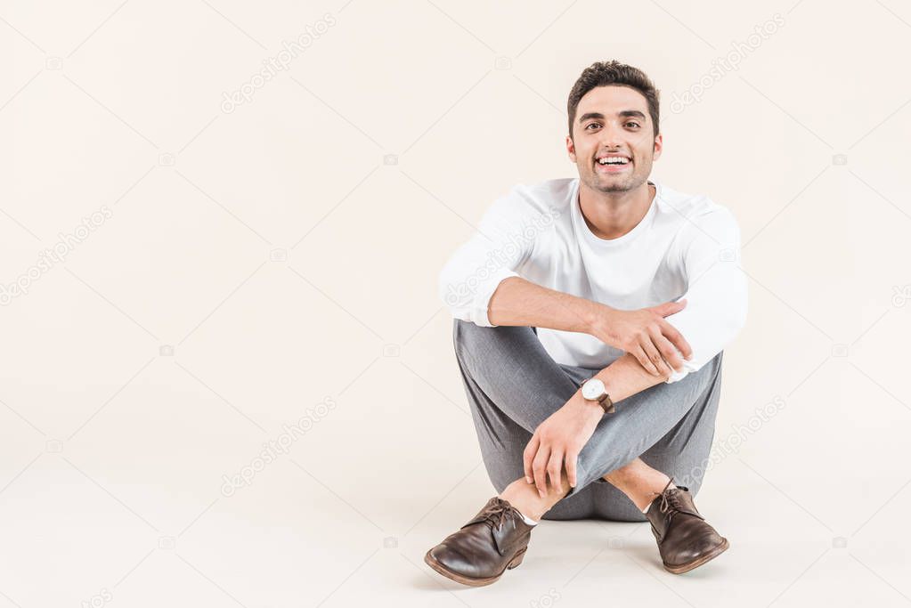 cheerful young man sitting and smiling at camera isolated on beige