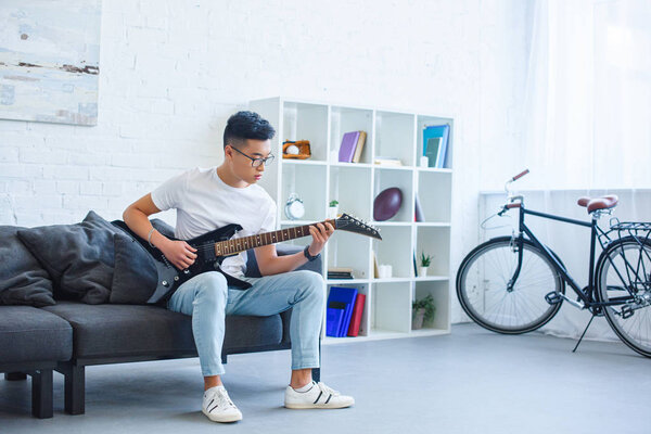 handsome asian man playing black electric guitar on sofa at home