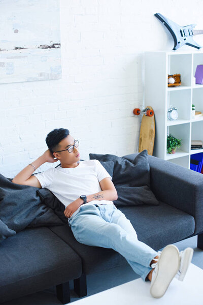 high angle view of pensive handsome asian man sitting on sofa with legs on table at home, looking away