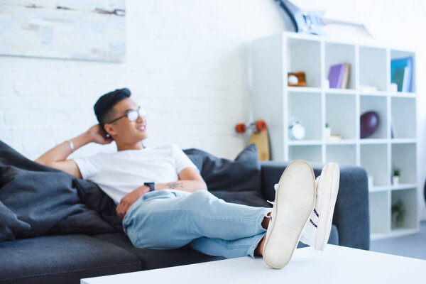 selective focus of handsome asian man sitting on sofa with legs on table at home, looking away
