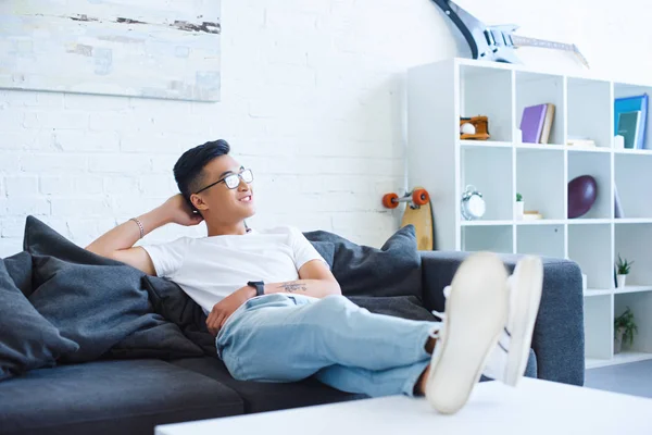 Smiling handsome asian man sitting on sofa with legs on table at home, looking away — Stock Image Smiling Handsome Asian Man Sitting Sofa Legs Table Home Looking Stock Image