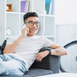 Leende stilig asiatisk man liggande på soffan och prata genom smartphone hemma