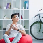 Handsome asian man sitting on red bean bag chair with cup of coffee and looking away at home