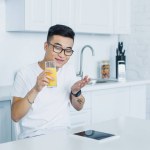 Happy young asian man in eyeglasses holding glass of juice and using digital tablet in kitchen