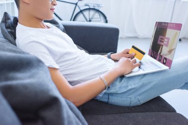 online bilet cihaz ile dizüstü bilgisayar kullanarak ve kredi kartı evde kanepenin üzerinde tutan adamın kırpılmış görüntü