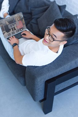 yakışıklı Asya adam kanepede yatan ve dizüstü online rezervasyon ile evde parçalar yüksek açılı görünüş