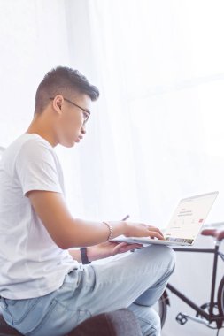 Asya yakışıklı kanepe evde sayfasında yüklü airbnb ile dizüstü bilgisayar kullanarak yan görünüm