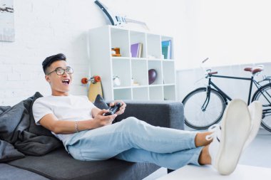 excited handsome asian man playing video game on sofa in living room clipart