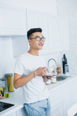 handsome smiling young asian man in eyeglasses holding cup of coffee and looking away in kitchen clipart