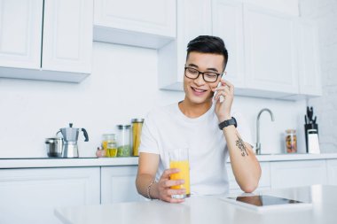 Genç Asyalı adam bardak suyu tutarak ve mutfakta otururken smartphone ile konuşurken gözlük gülümseyen
