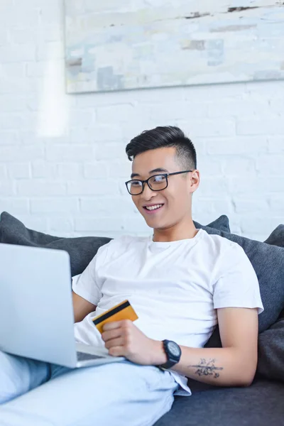 Sorrindo Bonito Asiático Homem Usando Laptop Sofá Segurando Cartão Crédito — Fotografia de Stock