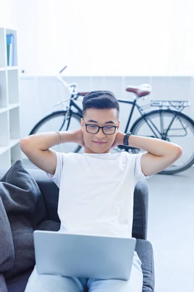 Весёлый Красивый Азиатский Мужчина Лежит Руками Головой Диване Смотрит Ноутбук — Бесплатное стоковое фото