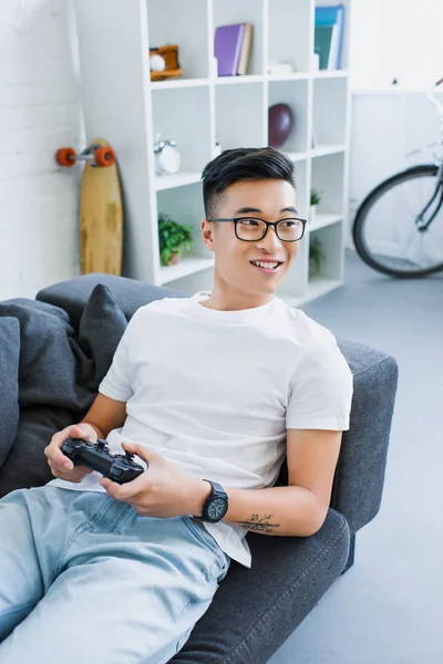 Sorrindo Bonito Asiático Homem Jogar Vídeo Game Deitado Sofá Casa — Fotografia de Stock