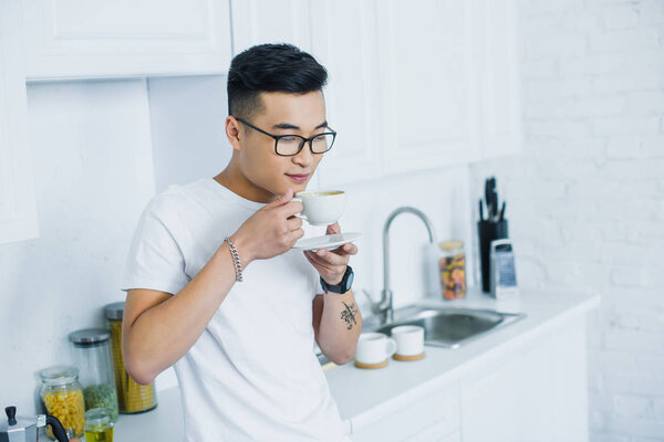 Красивый молодой азиат в очках держит чашку кофе на кухне
