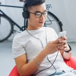 Leende ung asiatisk man i hörlurar med smartphone medan du sitter i böna väska stol hemma