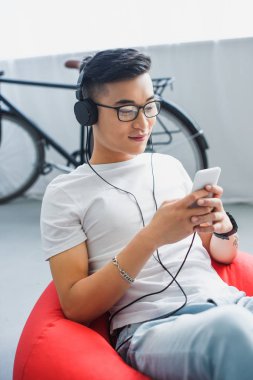 Genç Asyalı adam evde fasulye çanta sandalyede otururken smartphone kullanarak Kulaklığımda gülümseyen 