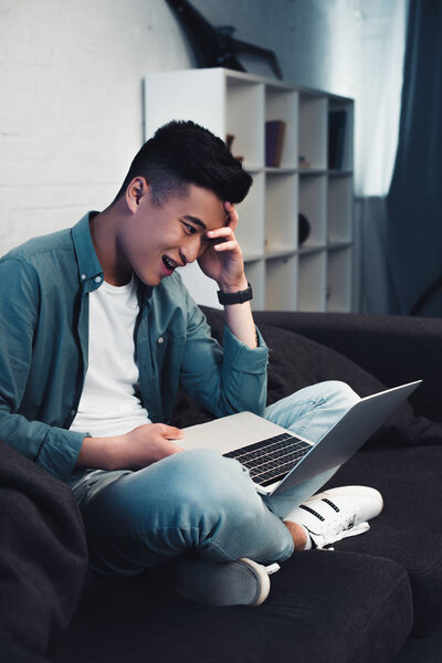 smiling young asian man sitting on couch and using laptop at home